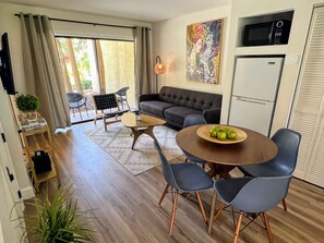 Una sala de estar moderna con un sofá, una mesa de comedor con sillas, una zona de cocina con microondas y un amplio ventanal con cortinas.