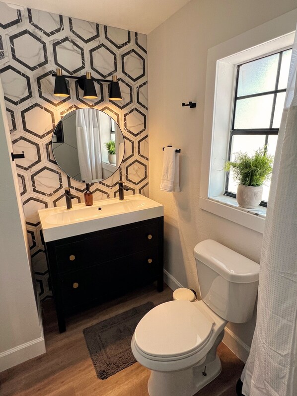 Un baño con pared de patrón hexagonal, un espejo redondo, un lavabo blanco con gabinete negro, un inodoro blanco y una ventana con planta en maceta.