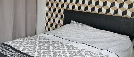 A bedroom with a geometric patterned wall, a bed with a patterned bedspread, and a curtain on the window.