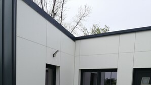 A modern outdoor patio with a wooden table and chairs, surrounded by a white building with large windows and a dark door. - New single-storey house 800 metres from the sea and footpaths (Pleubian)