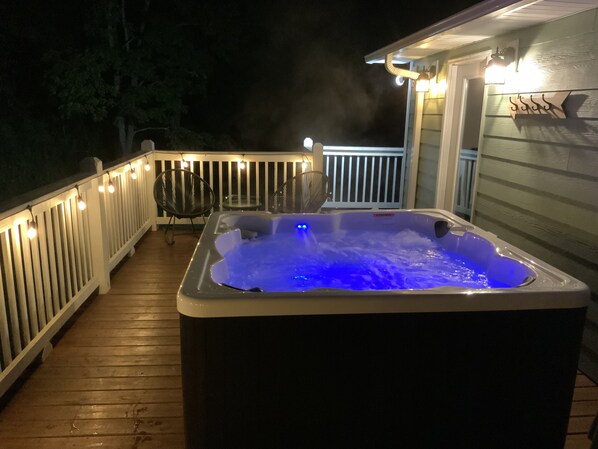A hot tub on a wooden deck with string lights, a chair, and a railing. - Hot Tub+Fire Pit, WiFi, Close to AVL+Burnsville! Perfect Location! Easy Access! (Burnsville)