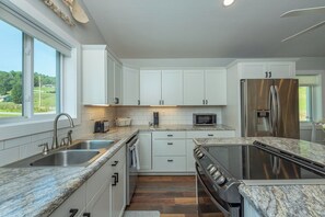 A modern kitchen with white cabinets, stainless steel appliances, and a large window offering a view of the outdoors. - Cozy Cottage~Hot Tub+Fire Pit+Grill~WiFi~Near AVL! (Burnsville)
