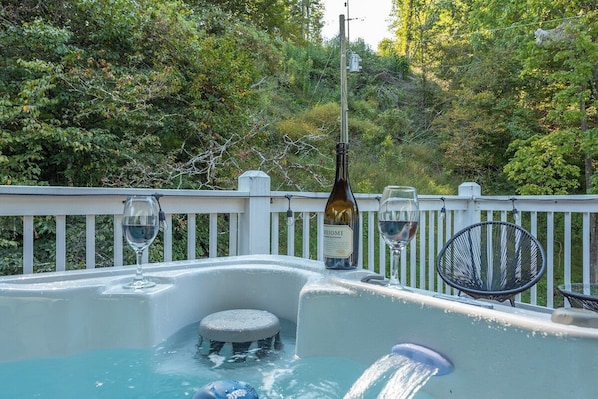 A hot tub with a bottle of wine and glasses, surrounded by a white fence and trees.
