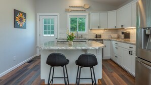 Een moderne keuken met witte kasten, een groot eiland met twee barkrukken, een roestvrijstalen koelkast en een raam met uitzicht op groen.