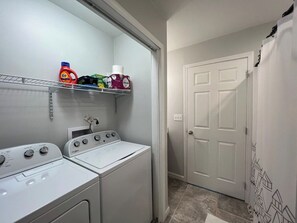 A laundry room with two white Whirlpool washers and dryers, a shelf with laundry supplies, and a white door with a geometric pattern. - Cozy Cottage~Hot Tub+Fire Pit+Grill~WiFi~Near AVL! (Burnsville)