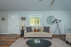 A living room with a beige sofa, a round coffee table, a lamp, a window, and a mirror on the wall. - Cozy Cottage~Hot Tub+Fire Pit+Grill~WiFi~Near AVL! (Burnsville)