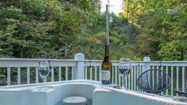 Een jacuzzi met een fles wijn en glazen, omgeven door een witte hek en weelderige begroeiing.