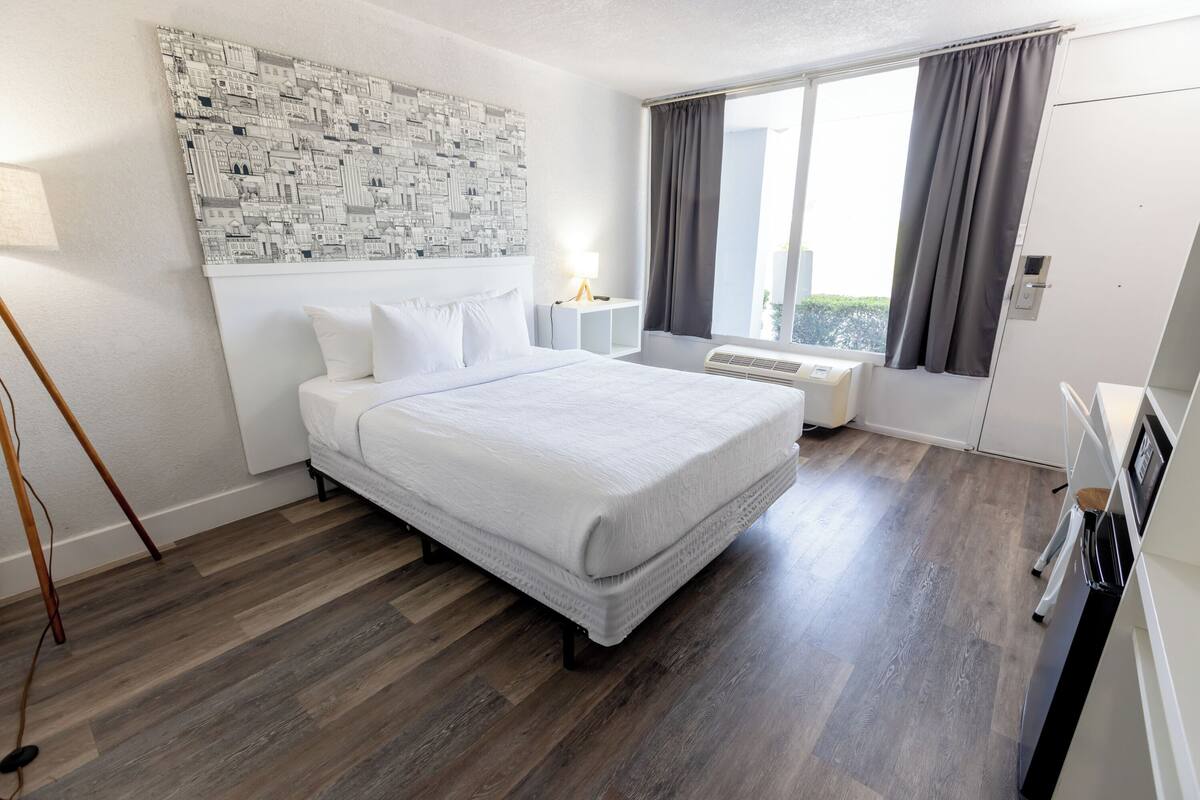 Hotellihuone, jossa on suuri sänky, pääty, jossa on kaupunkimaisemaa esittävä kuvio, yöpöytä lampulla, ikkuna verhoilla ja sisäänrakennettu ilmastointilaite.