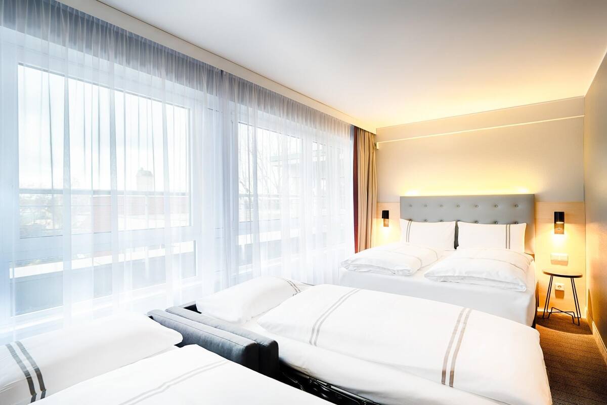 A hotel room with two beds, a large window with sheer curtains, a headboard with a light fixture, and a small bedside table with a lamp.