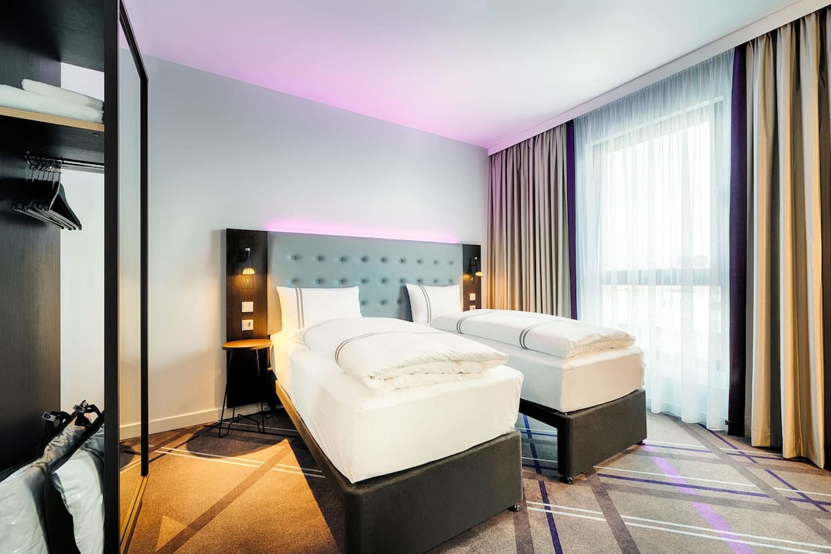 A hotel room with two beds, a headboard with a light panel, a small table, and a large window with curtains.