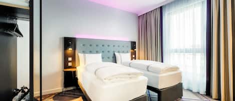 Habitación de hotel con dos camas, cabecera con panel de luz, una mesita y un ventanal con cortinas.