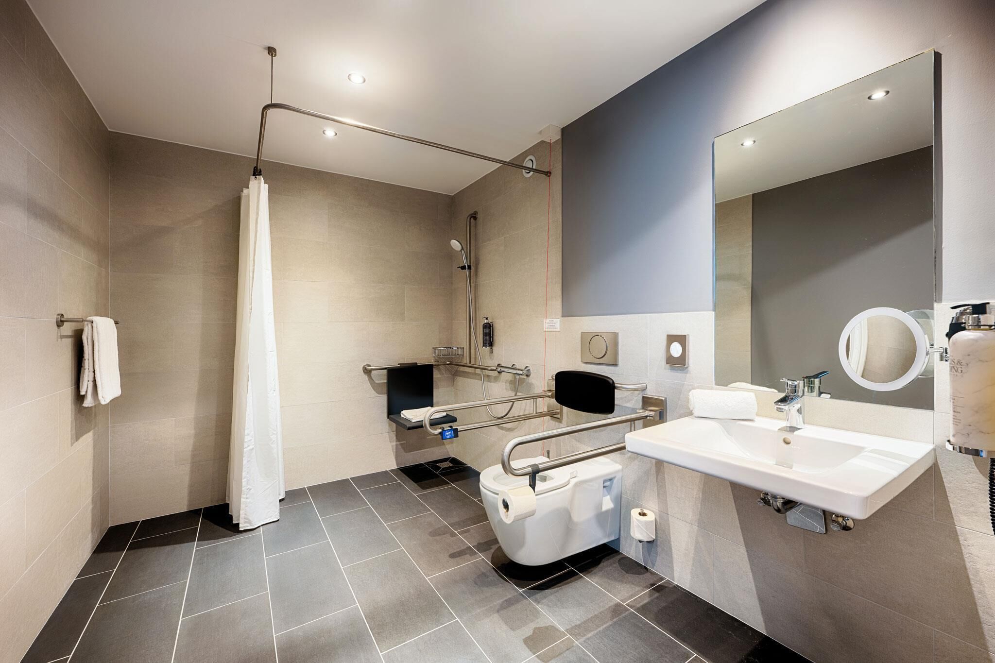 A modern bathroom with a shower, toilet, and sink, equipped with grab bars and a hand dryer.