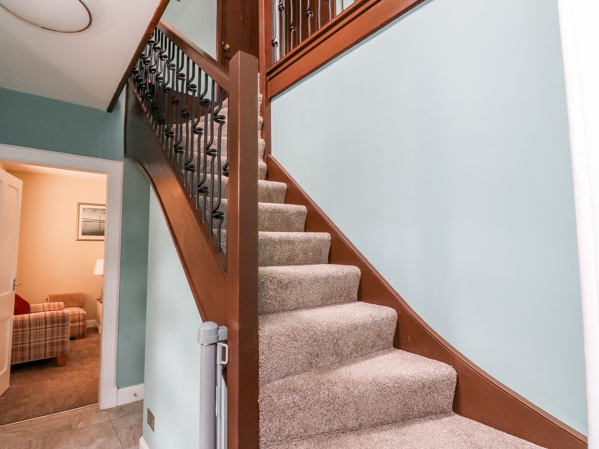 Una escalera alfombrada con barandilla de madera y balaustrada de metal negro, que conduce a una habitación con pared azul claro y un cuadro enmarcado.