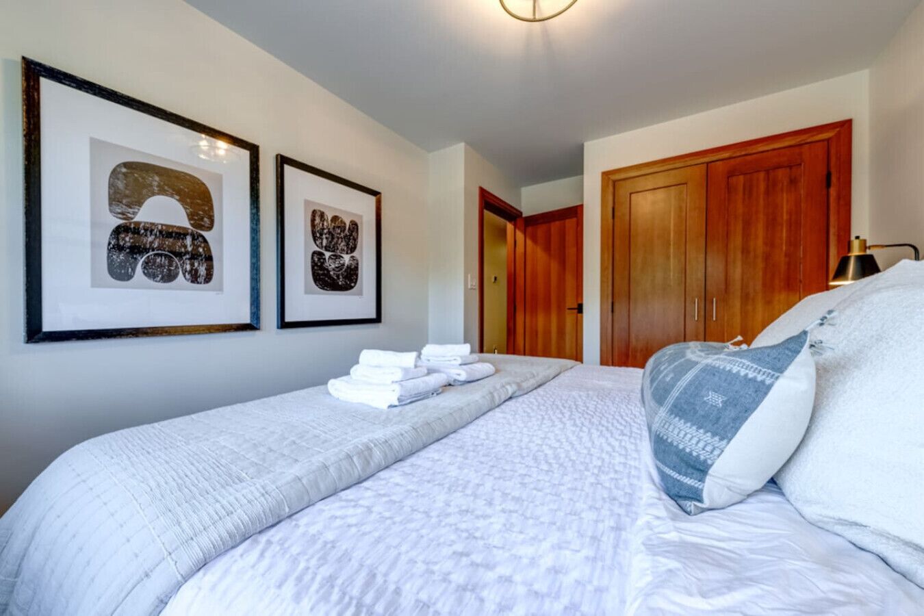 A bedroom with a bed, two framed pictures, a wooden wardrobe, and a bedside lamp.