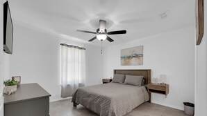 Ein Schlafzimmer mit Bett, Nachttischen, Deckenventilator, Wandlampe, einem Bild, einem Fenster mit Vorhängen und einer kleinen Mülltonne.