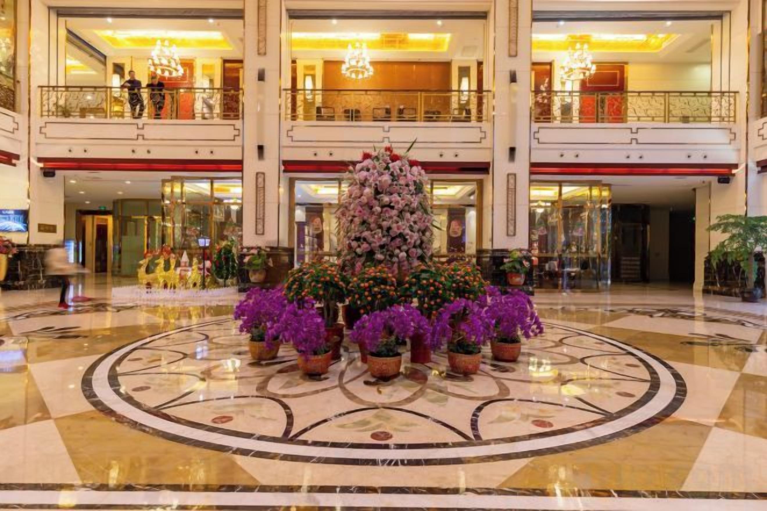 A hotel lobby with a central floral arrangement, a grand staircase, and a chandelier.