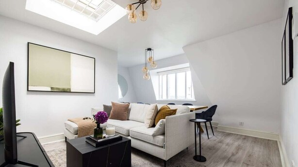 Ein modernes Wohnzimmer mit einem weißen Sofa, einem schwarzen Couchtisch und einem Fernseher. Über dem Sofa befindet sich ein Oberlicht und zur Rechten ein Fenster.