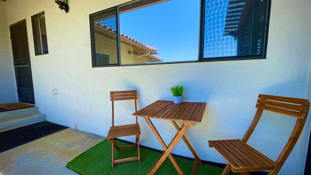 A patio with a wooden table and two chairs, a small potted plant, and a window overlooking a building.