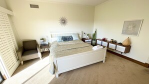 A bedroom with a white bed, a bedside table, a chair, a nightstand, a wall-mounted shelf, and a decorative wall piece.