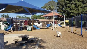 A playground with slides and swings, covered by large umbrellas, and surrounded by trees.