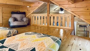 Une chambre confortable située dans les combles, avec un plafond en bois, un fauteuil rembourré, un lit recouvert d’une courtepointe et un balcon orné d’une pancarte sur laquelle est écrit « BIENVENUE ».