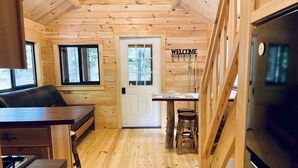 L’intérieur d’une cabane en bois comprend un coin cuisine, une table à manger avec des tabourets et une pancarte « BIENVENUE » sur la porte.