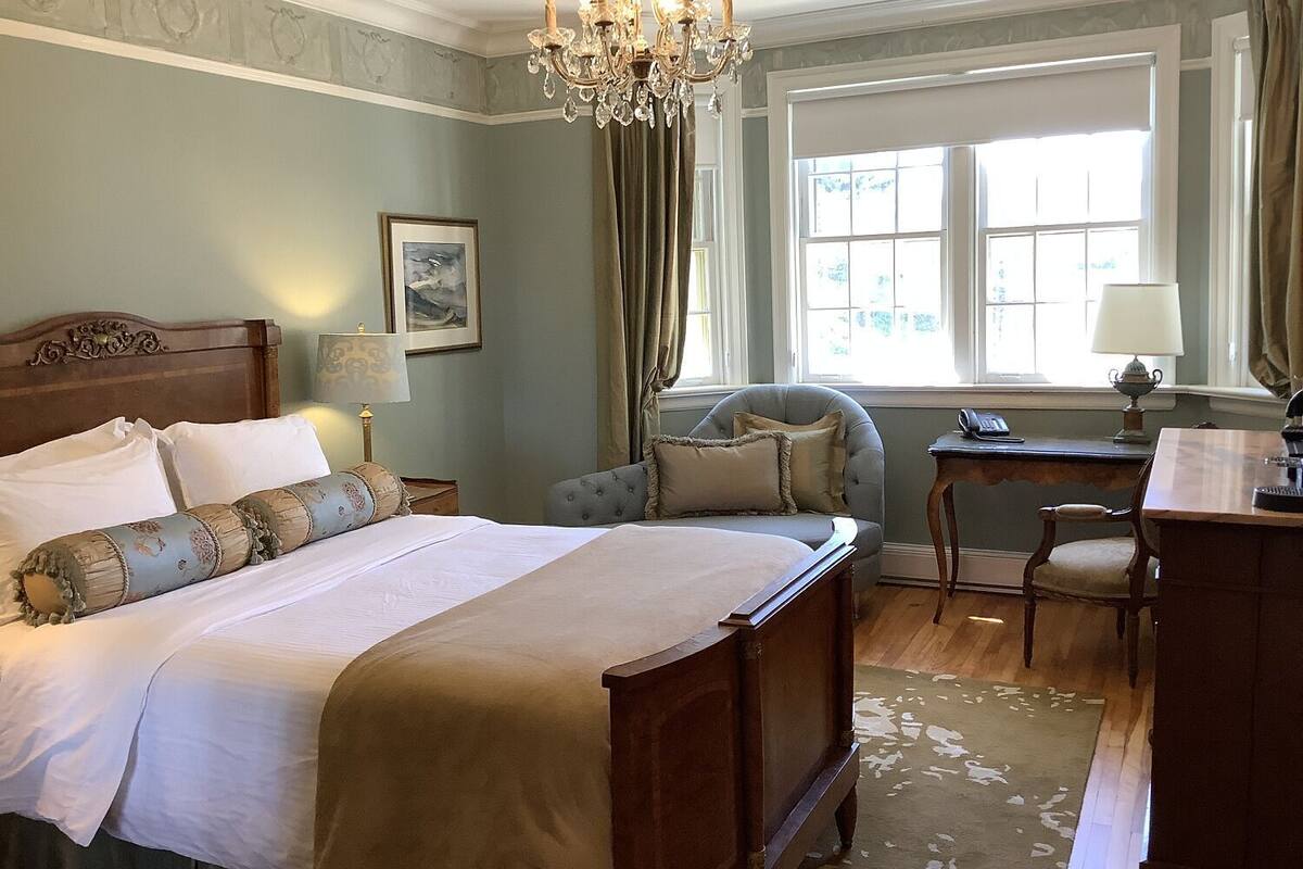 A bedroom with a large bed, a desk, a chair, a chandelier, and a window with curtains.