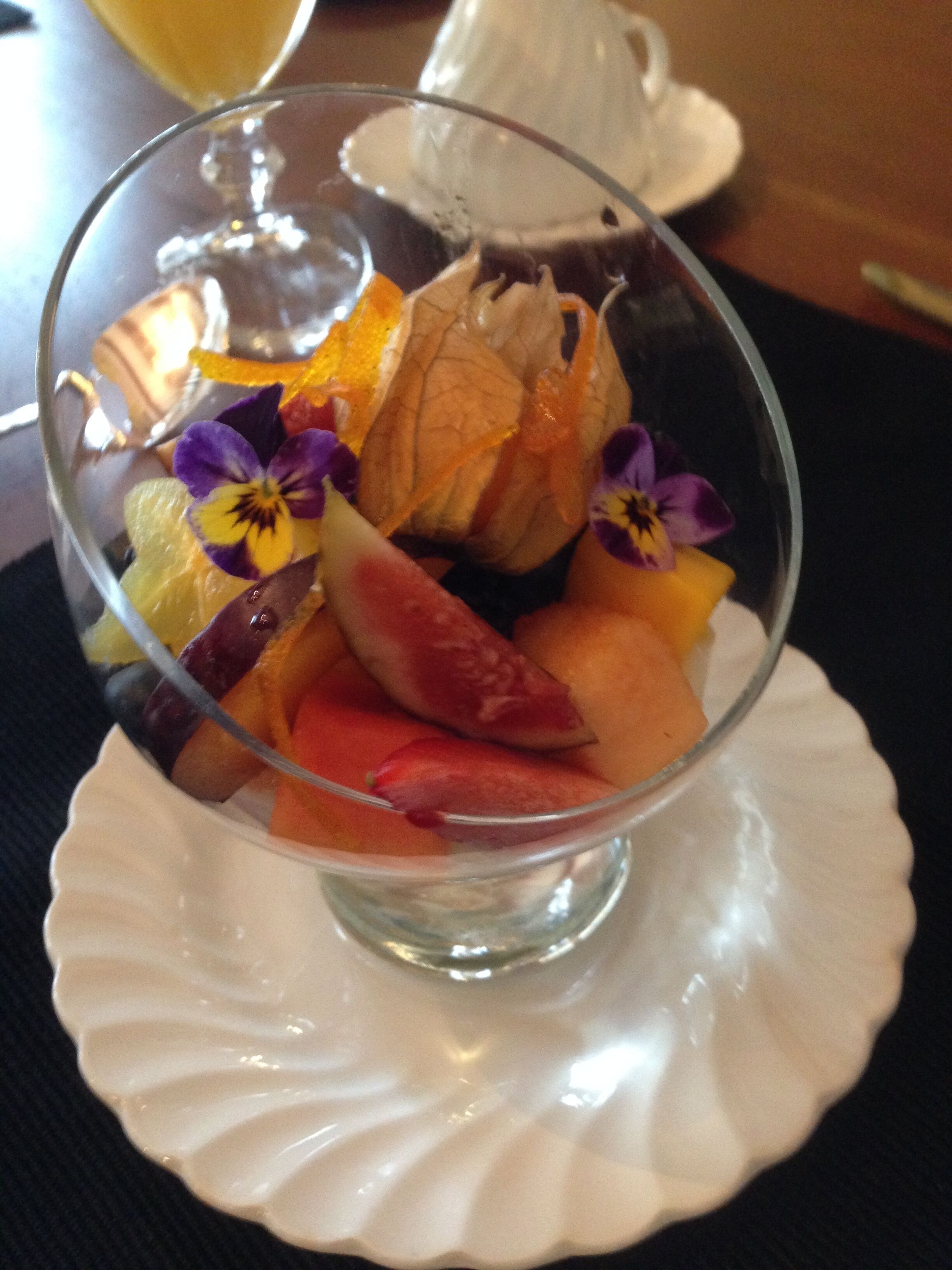 Eine transparente Glasschale mit Obstsalat, bestehend aus verschiedenen Früchten und Blüten, auf einem weißen Teller mit gewelltem Rand.
