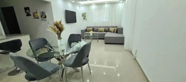 A modern living room with a grey sofa, a glass dining table with four chairs, and a flat-screen TV mounted on the wall.
