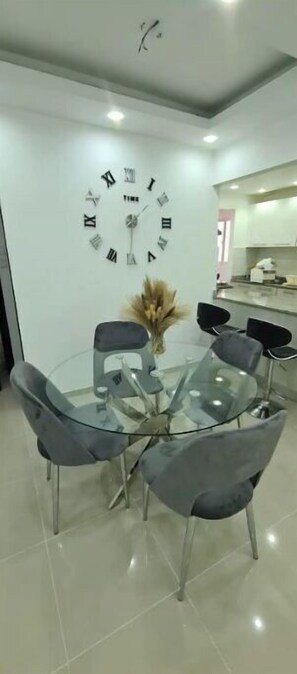 A modern dining area with a round glass table surrounded by six chairs, a wall-mounted clock, and a kitchen in the background.