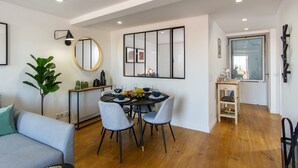 Un coin repas avec une table ronde, deux chaises et un miroir au-dessus. On y trouve une plante et une petite table sur laquelle sont posés divers objets.