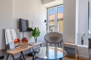 Un salon moderne avec un téléviseur, une table en verre, un fauteuil en osier et une vue sur un immeuble dont le toit est recouvert de tuiles.