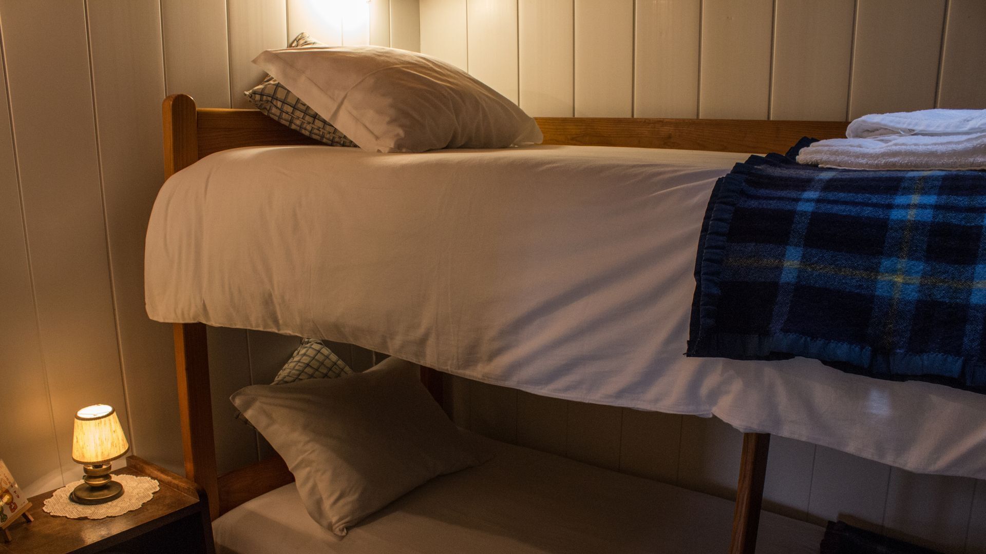 Een stapelbed met een deken en kussens, een bedlampje en een klein tafeltje met een placemat.