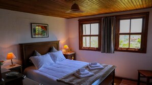 Een net opgemaakt bed met witte lakens, een houten hoofdbord en een raam met gordijnen. Er staan twee bedlampen en een ingelijste foto aan de muur.