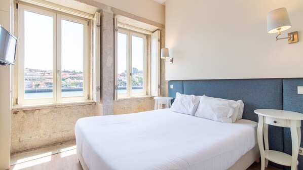 A hotel room with a bed, a small table, a chair, a wall-mounted lamp, and large windows with a city view. - Blue Oporto Home River Front - Ribeira (Vila Nova de Gaia)