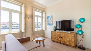 Uma sala de estar com um sofá, uma mesinha redonda, um armário de madeira com uma TV e vista para a paisagem urbana através de grandes janelas.