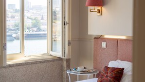 Een hotelkamer met uitzicht op een rivier, een bed met een gestreept hoofdeinde, een klein tafeltje met een kopje en glazen, en een wandlamp.