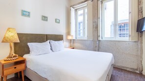 Un lit avec du linge de lit blanc, une table de chevet en bois avec une lampe et un téléviseur fixé au mur.