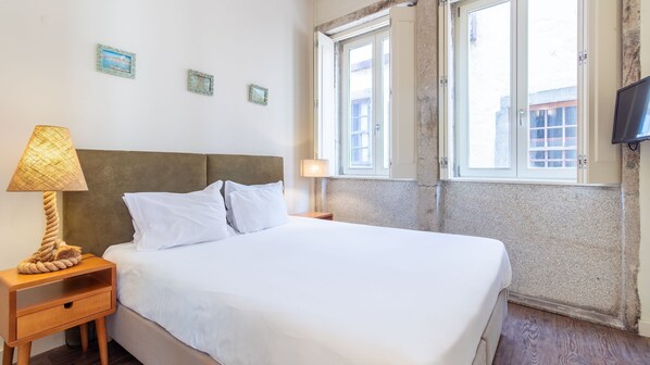 A bed with white linens, a wooden nightstand with a lamp, and a television on the wall.