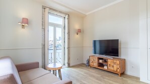 A modern living room with a sofa, a small round table, a TV on a wooden cabinet, and a city view through a large window. - Purple Oporto Home River Front - Ribeira (Vila Nova de Gaia)
