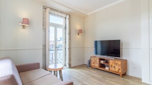 A modern living room with a sofa, a small round table, a TV on a wooden cabinet, and a city view through a large window. - Purple Oporto Home River Front - Ribeira (Vila Nova de Gaia)
