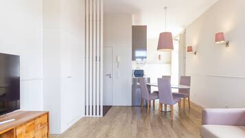 Uma sala de estar moderna com televisão de tela plana, um móvel para TV de madeira, uma área de refeições com mesa e cadeiras e uma cozinha ao fundo.