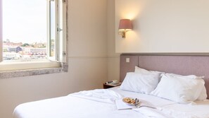 A hotel room with a bed, a nightstand, a lamp, and a window with a view of buildings. - Purple Oporto Home River Front - Ribeira (Vila Nova de Gaia)