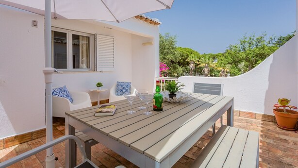 Um pátio externo com mesa de madeira, cadeiras brancas e uma pequena mesa redonda com uma planta.