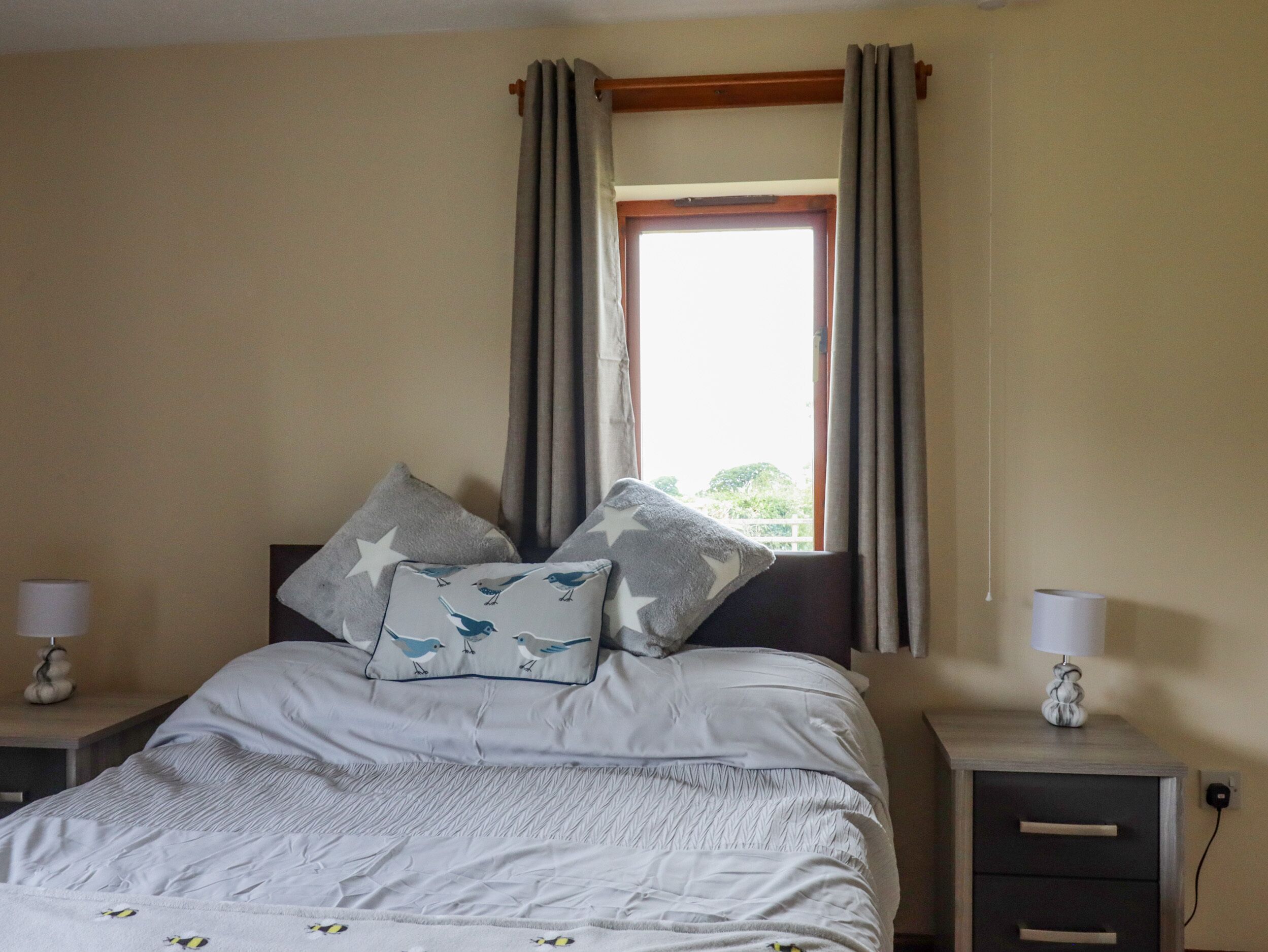A bedroom with a bed, two nightstands, a window with curtains, and a view of greenery outside.
