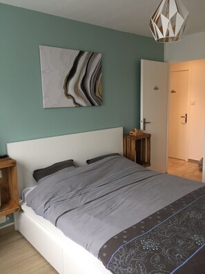 Een slaapkamer met een bed, een nachtkastje, een abstract schilderij aan de muur, een deur en een hanglamp.