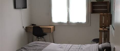 A bedroom with a bed, a desk, a chair, a TV, and a window.