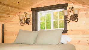 Un dormitorio de cabaña de madera con cama, dos lámparas fijadas a la pared y ventana con vista a la vegetación.