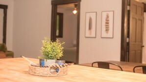 Una mesa de comedor con una cesta tejida que contiene una planta y velas, una silla de madera y cuadros en la pared.