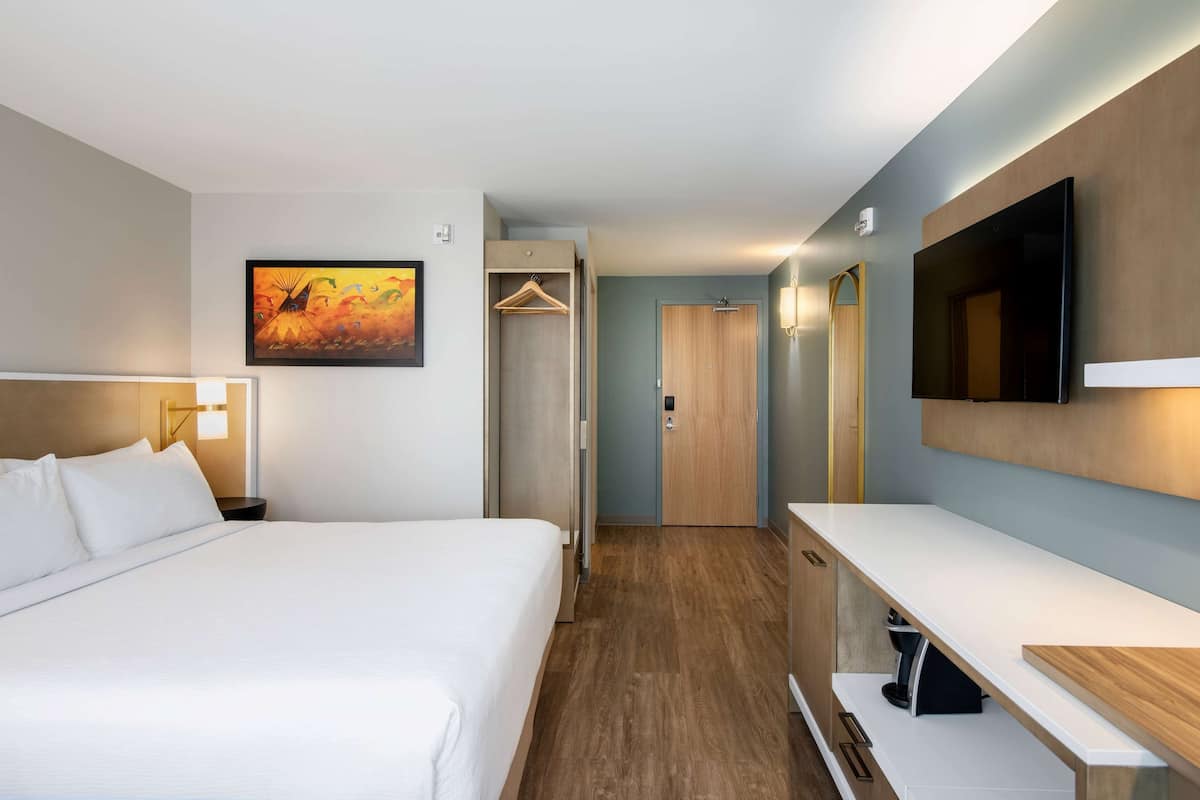 Habitación de hotel con cama, mesita de noche, televisor, perchero y puerta que da al baño.